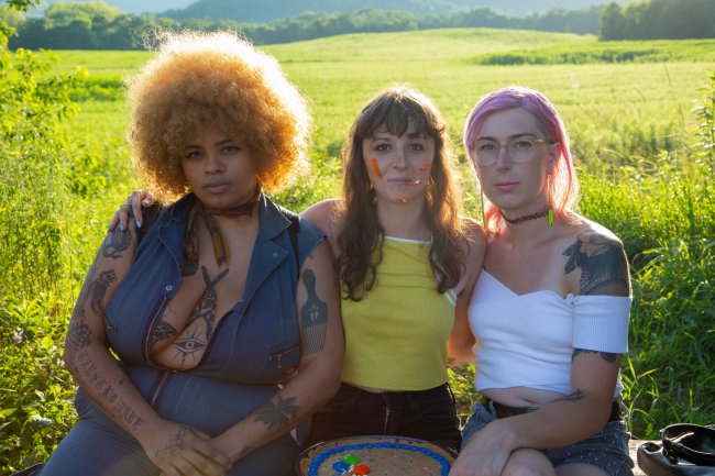Three artists pose arm-in-arm in front of a green pasture at sunset